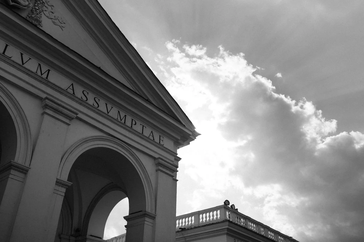 Black and white photo of classical building with large archways, columns, and Latin inscription, against cloudy sky.