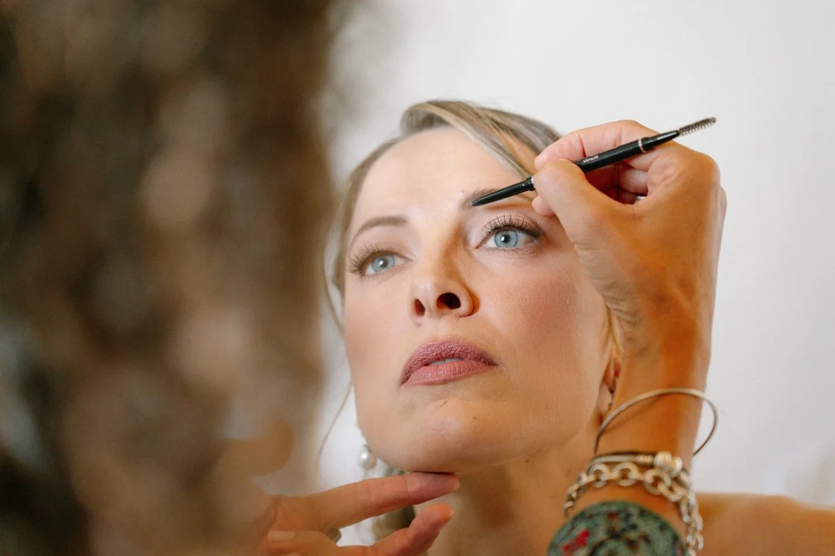 A woman with blonde hair and blue eyes having her makeup done, with a makeup artist applying eyeliner or eyeshadow on her eyelid.