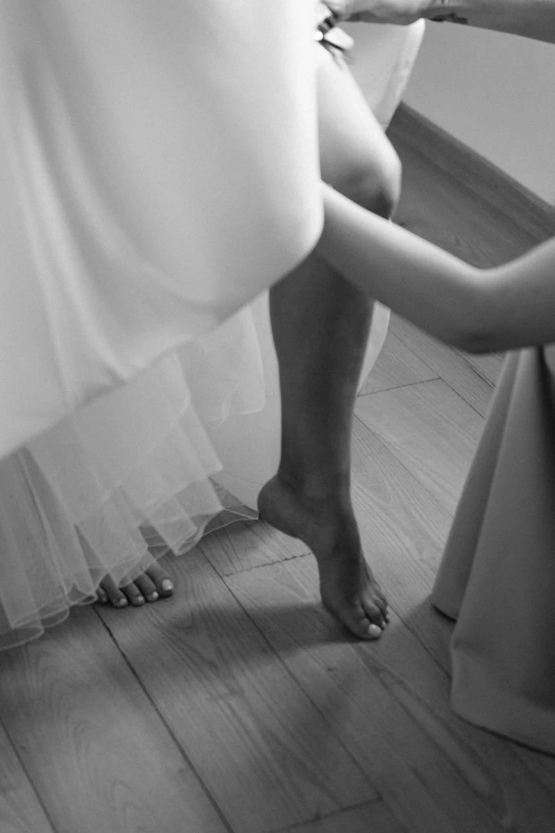 Foot of a bride in a wedding dress, standing on a wooden floor, with a groom's hand touching her waist.