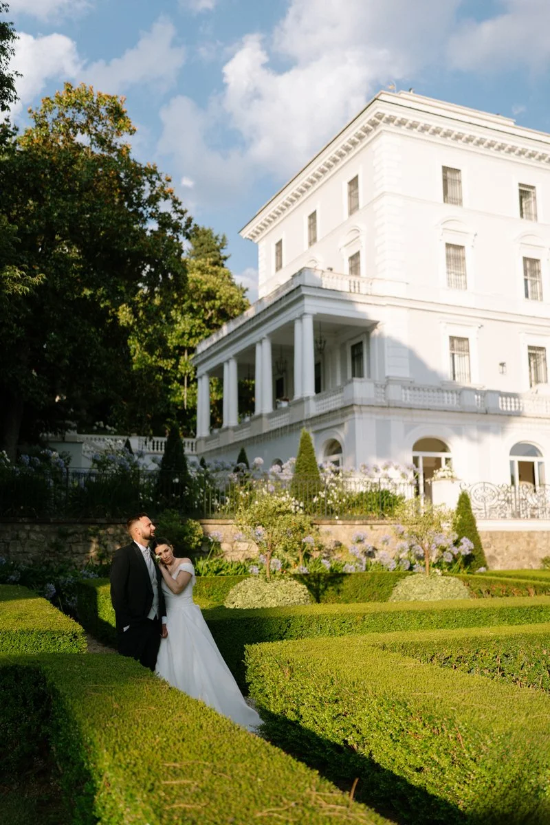 A newlywed couple posing in front of a white mansion with well-manicured gardens, green hedges, and flowers under a partly cloudy sky.