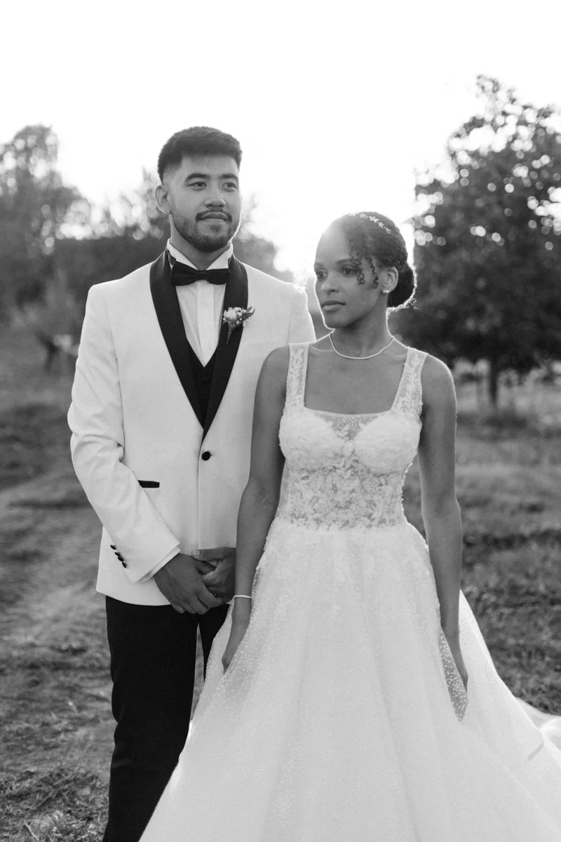 A black and white photo of a bride and groom outdoors, with the groom in a white tuxedo and the bride in a lace wedding gown.