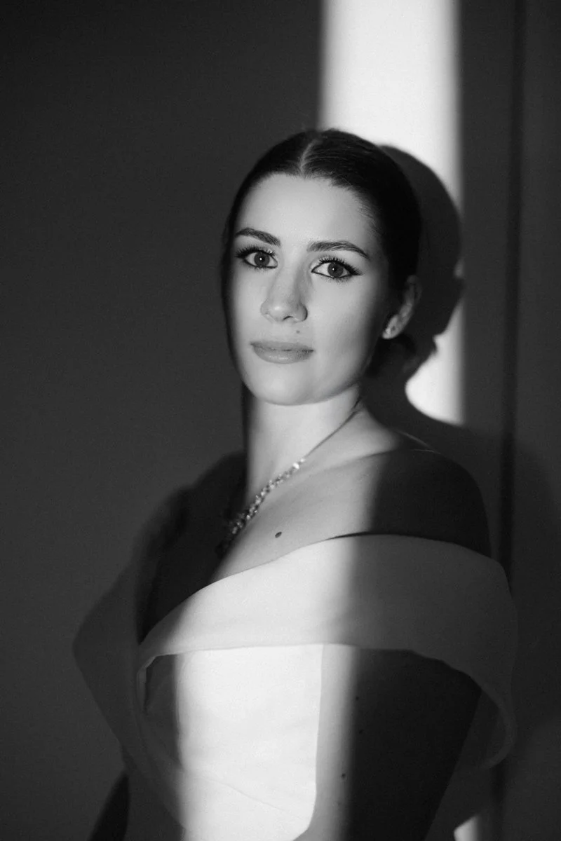 Black and white portrait of a woman with dark hair styled back, wearing a one-shoulder dress and a necklace, with light falling on her face and shoulder, creating shadows on her cheek and neck.