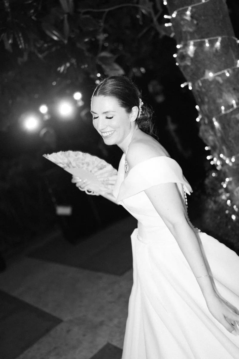 A woman in a wedding dress smiling while holding a fan at a celebration or wedding reception.