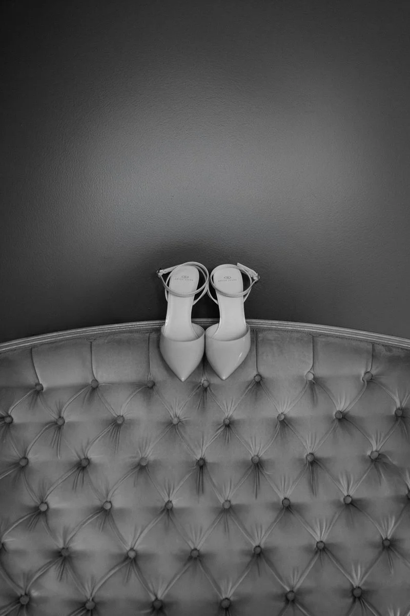 A pair of white high-heeled shoes placed upside down on a tufted, velvet-upholstered bench against a plain wall.