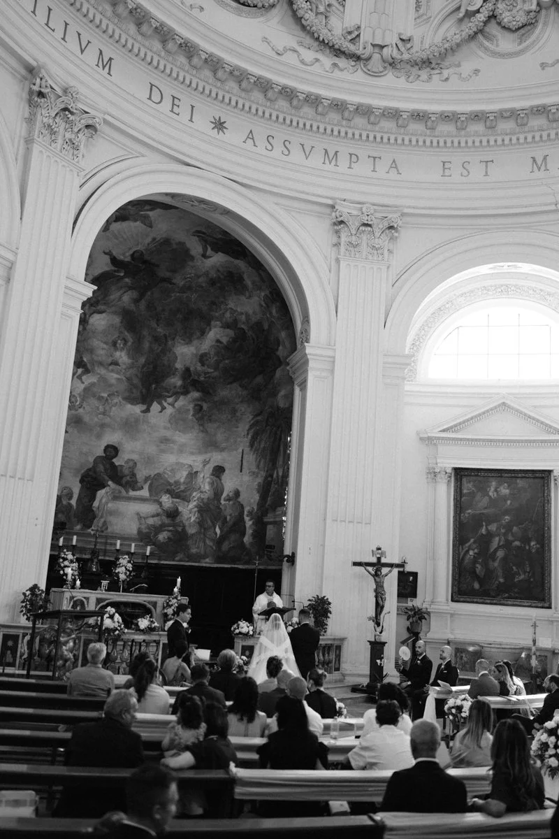 A wedding ceremony inside a grand church with ornate architecture and religious artwork, including a large painting and a crucifix, with guests seated facing the altar.