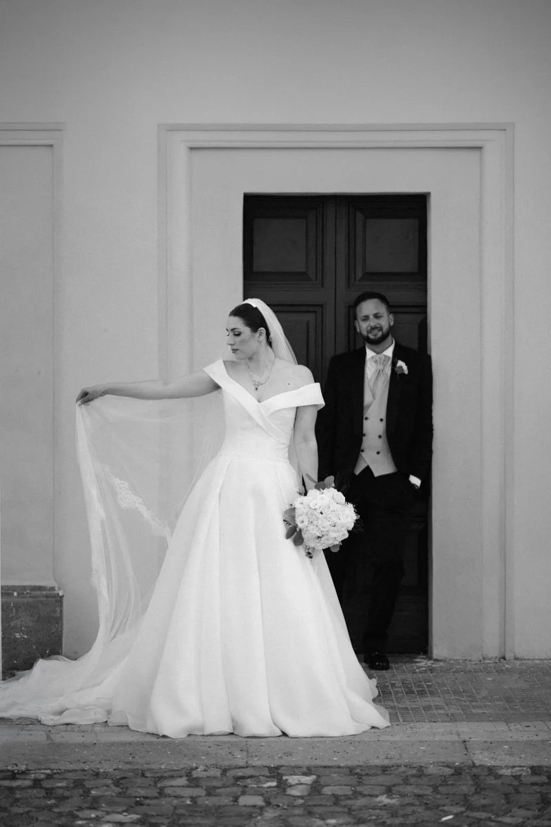 Black-and-white photo of a bride and groom standing at a doorway, with the bride holding a bouquet of flowers.