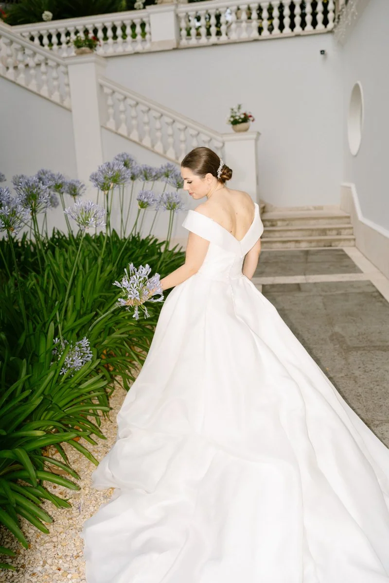 A bride in a white wedding dress standing outdoors near purple flowering plants, with a white building and staircase in the background.