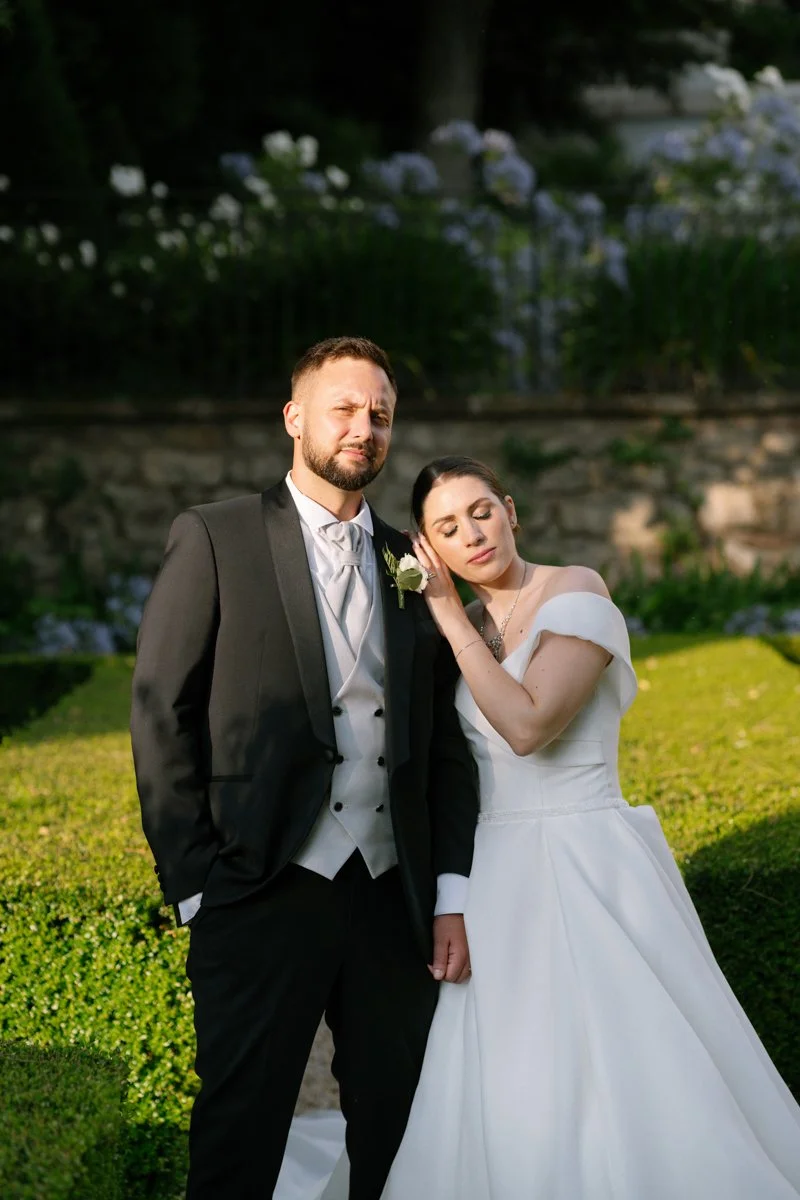 A bride and groom stand together outdoors in a garden, with the bride resting her head on the groom's shoulder. The groom is dressed in a black tuxedo with a white shirt and tie, while the bride is in a white off-shoulder wedding gown.