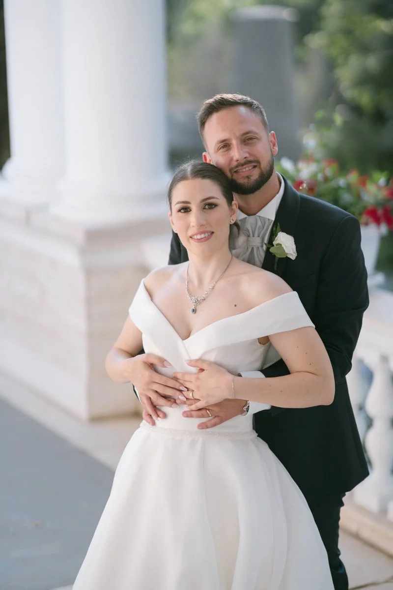 A newlywed couple in wedding attire, with the bride in a white off-the-shoulder gown and the groom in a dark tuxedo, standing outdoors in front of a white pillar and greenery.