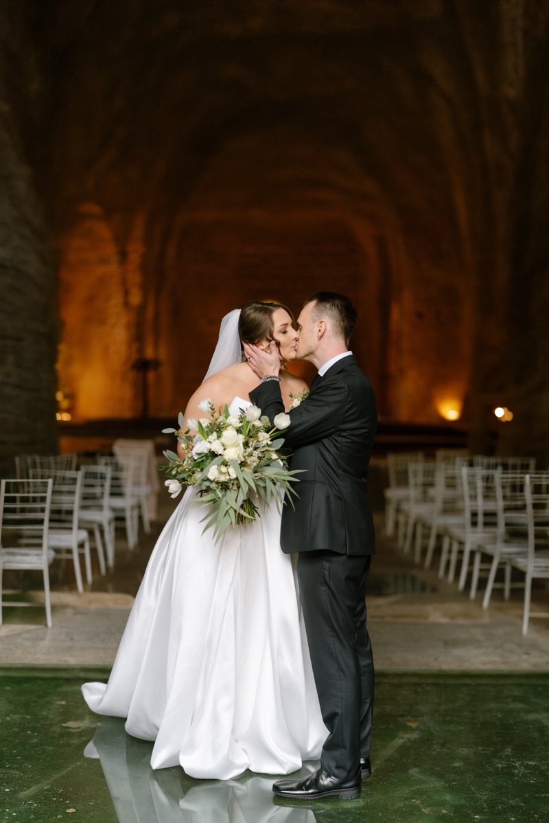 A bride and groom kiss during their wedding ceremony inside a grand, arched stone hall with seating on both sides. The bride holds a large bouquet of white flowers and greenery, and is wearing a strapless white wedding gown and veil. The groom is dre