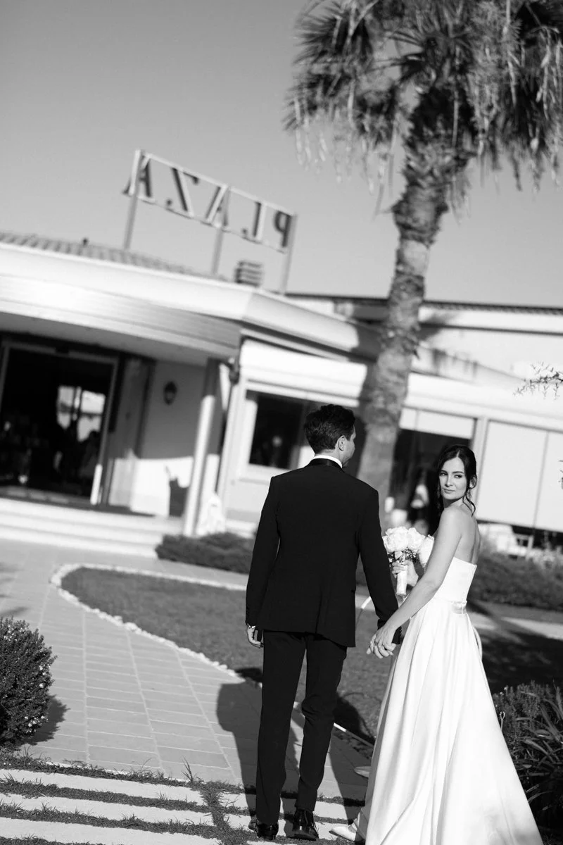 A black and white photo of a bride and groom holding hands outside a building with a palm tree. The bride is wearing a strapless wedding dress and holding a bouquet of flowers, and the groom is wearing a suit. The building has a sign with the word 'P