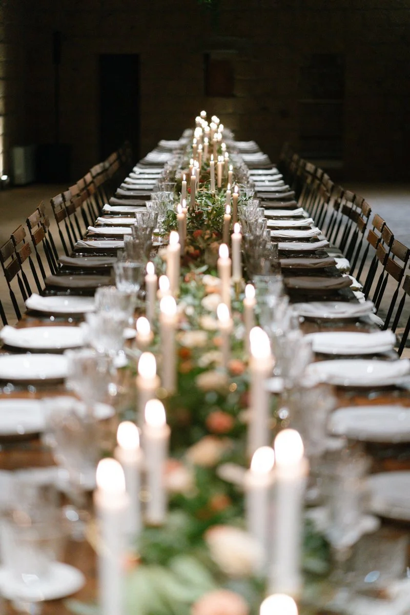 Long dining table decorated with candles, greenery, and flowers, set for a formal event or celebration in a dimly lit room.