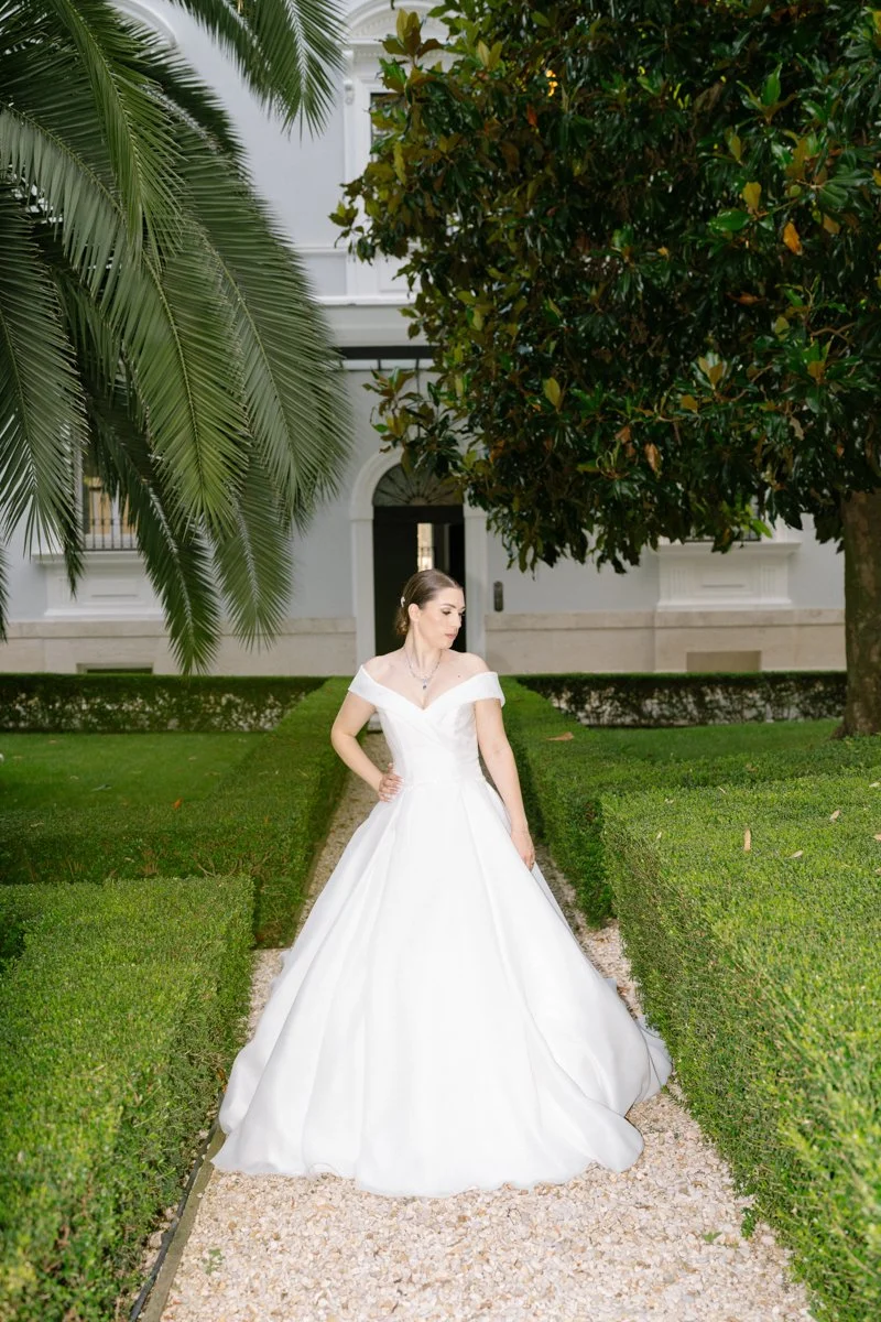 A bride in a white wedding dress standing on a gravel path between well-manicured hedges, with large green trees and a white building in the background.