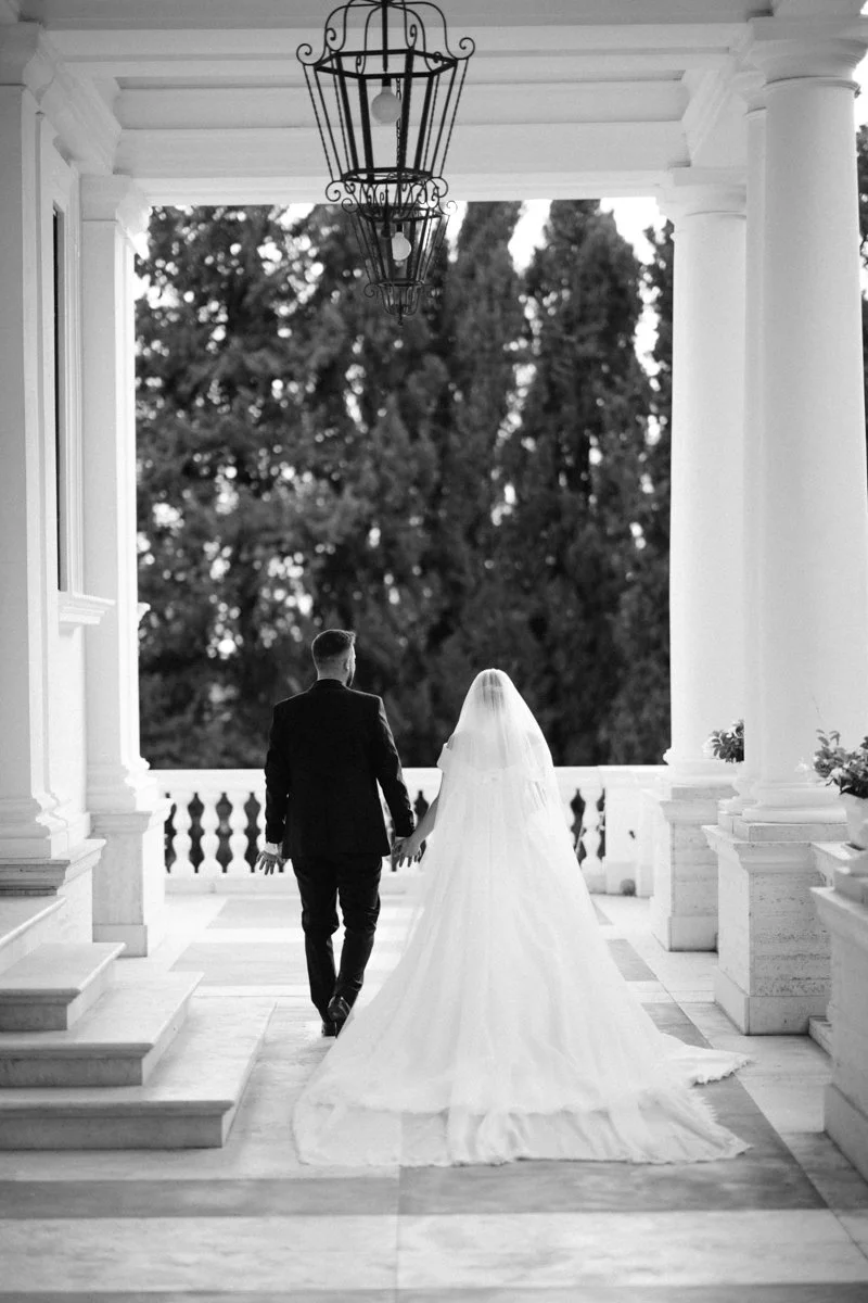 A bride and groom holding hands and walking on a porch with columns, surrounded by trees in the background.