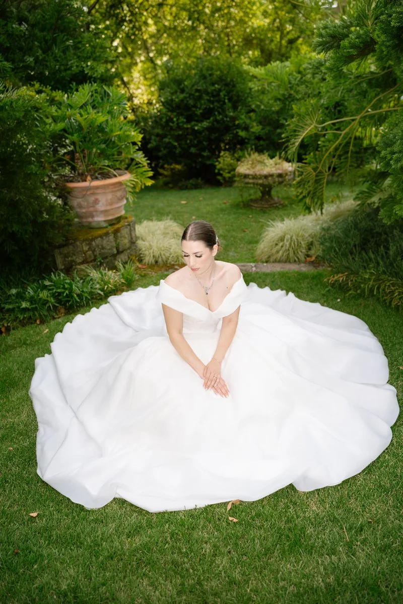 A woman in a white wedding dress sitting on the grass in a lush garden, surrounded by green plants and trees.