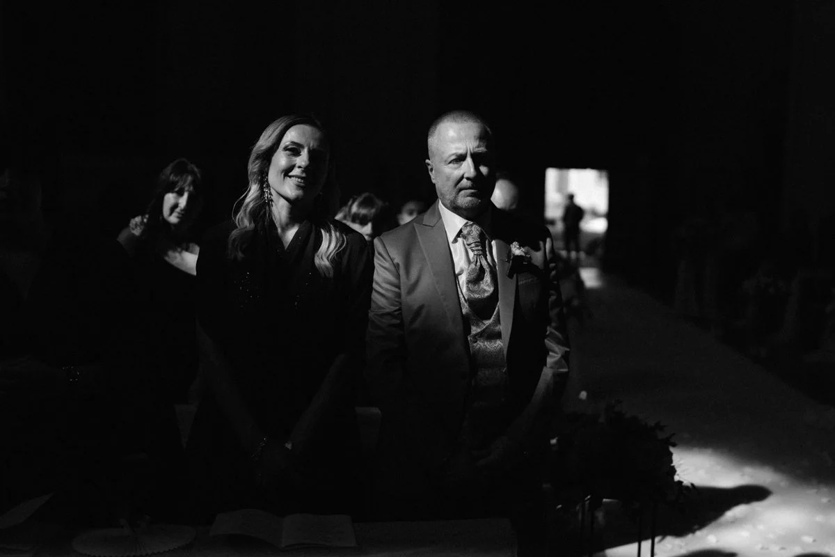 Black and white photo of a man and woman standing at a formal event, with other people blurred in the background.