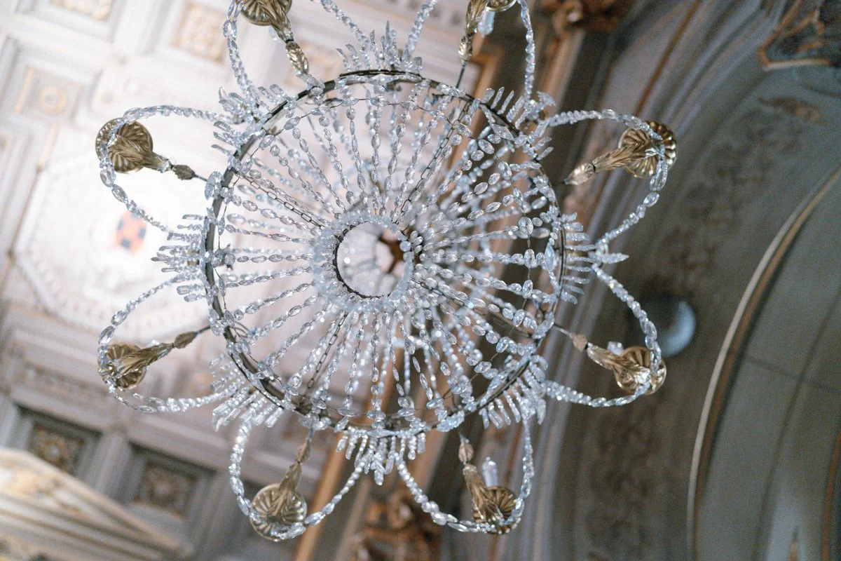 A large, ornate chandelier made of crystal and gold adorns a decorated ceiling, viewed from below.