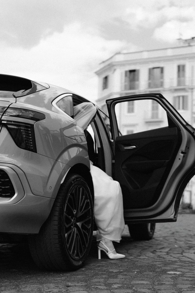 A person wearing white high heels and a long white dress is getting out of a modern car with the door open, parked on a cobblestone street with a large building in the background.