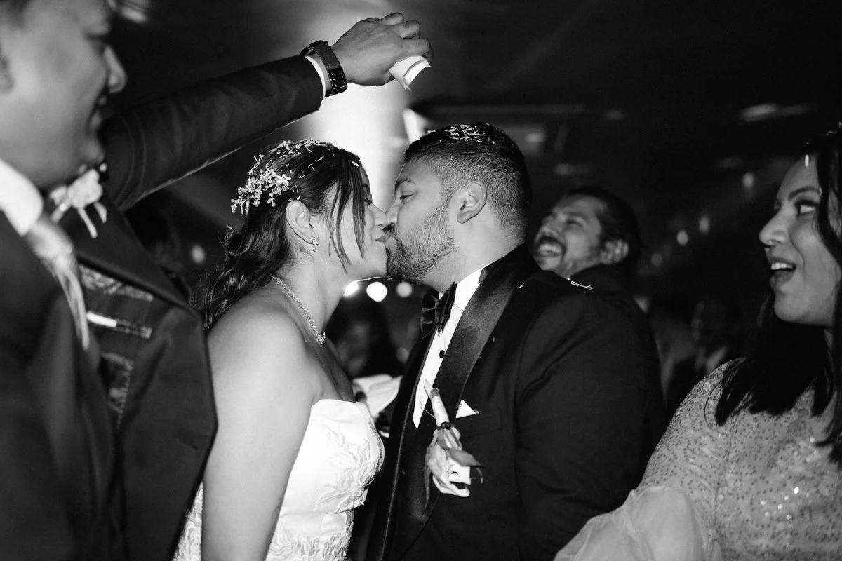 A black and white photo of a wedding celebration with a bride and groom sharing a kiss, surrounded by happy guests.