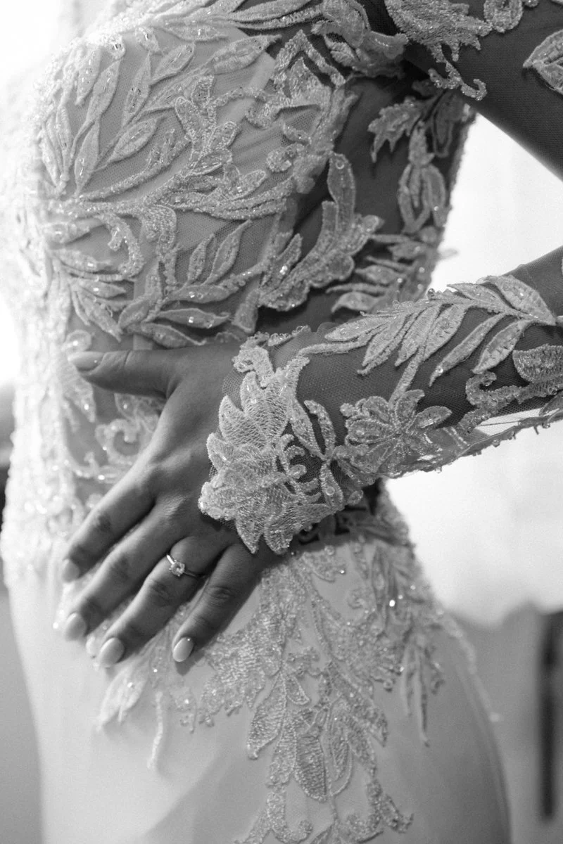 Close-up of a bride's hand with a ring, resting on her wedding dress with intricate lace and embroidery details.