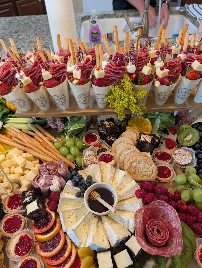 A spread of various desserts and fruits on a table, including cheese, cookies, grapes, strawberries, kiwi, and raspberry-topped pastries, with honey in the center for dipping.