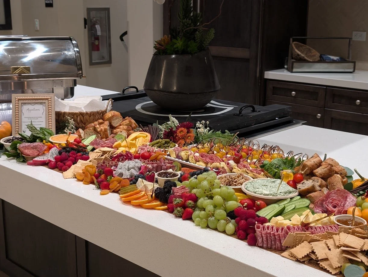 A long buffet table with an assortment of fruits, cheeses, meats, crackers, and snacks, decorated with flowers, set in a room with dark wood cabinets and a stove in the background.