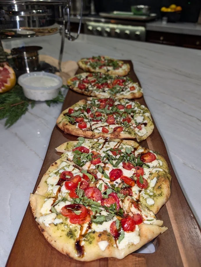 A wooden serving board with five slices of pizza topped with cherry tomatoes, basil, and drizzled with balsamic glaze, on a white marble countertop.