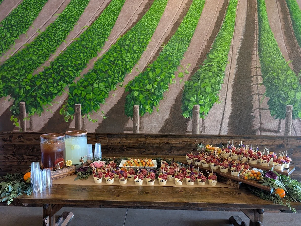 Table set with beverage dispenser, plastic cups, and small dessert cups topped with fruit, in front of a mural of green vineyard rows.