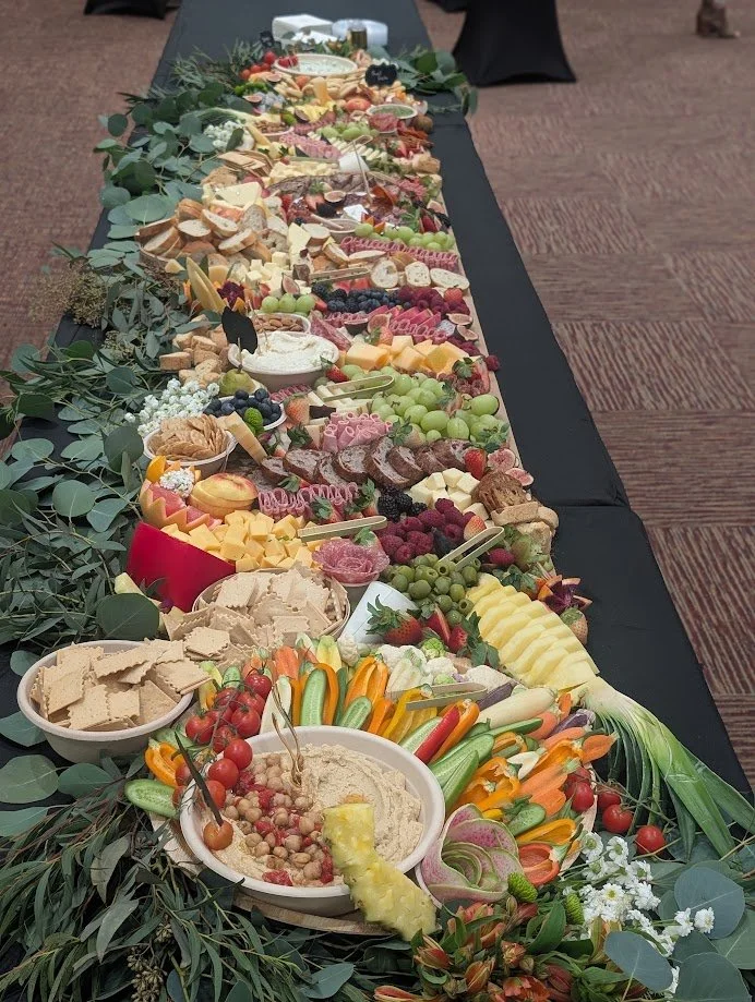 A long table decorated with a variety of cheeses, fruits, vegetables, crackers, and dips, arranged with greenery and flowers.