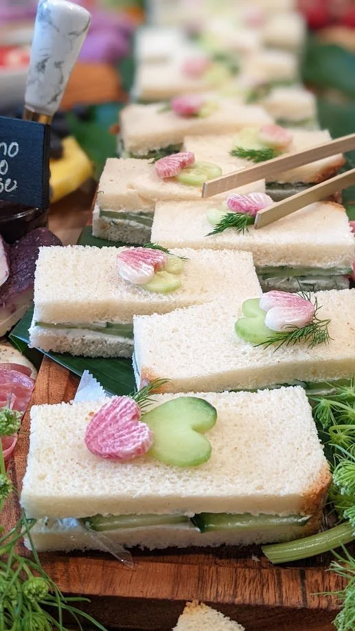 Close-up of finger sandwiches with cucumber slices and pink and white decorative garnishes on top, arranged on a wooden platter at a buffet or market.