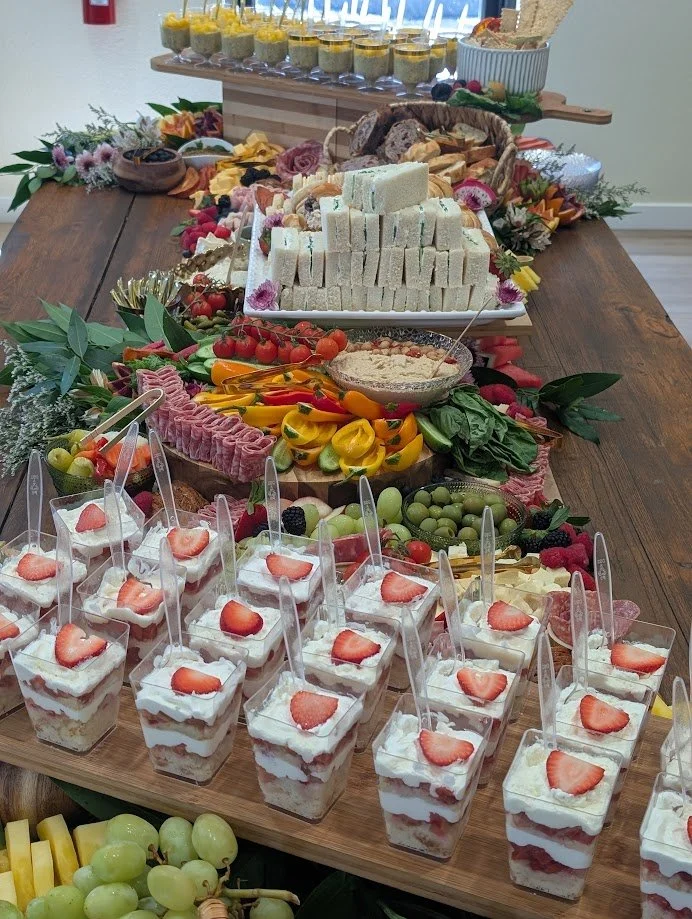 A dessert buffet with strawberry parfaits, sliced bread, cheese, assorted vegetables, fruits, dips, and small cups of layered desserts with strawberries.