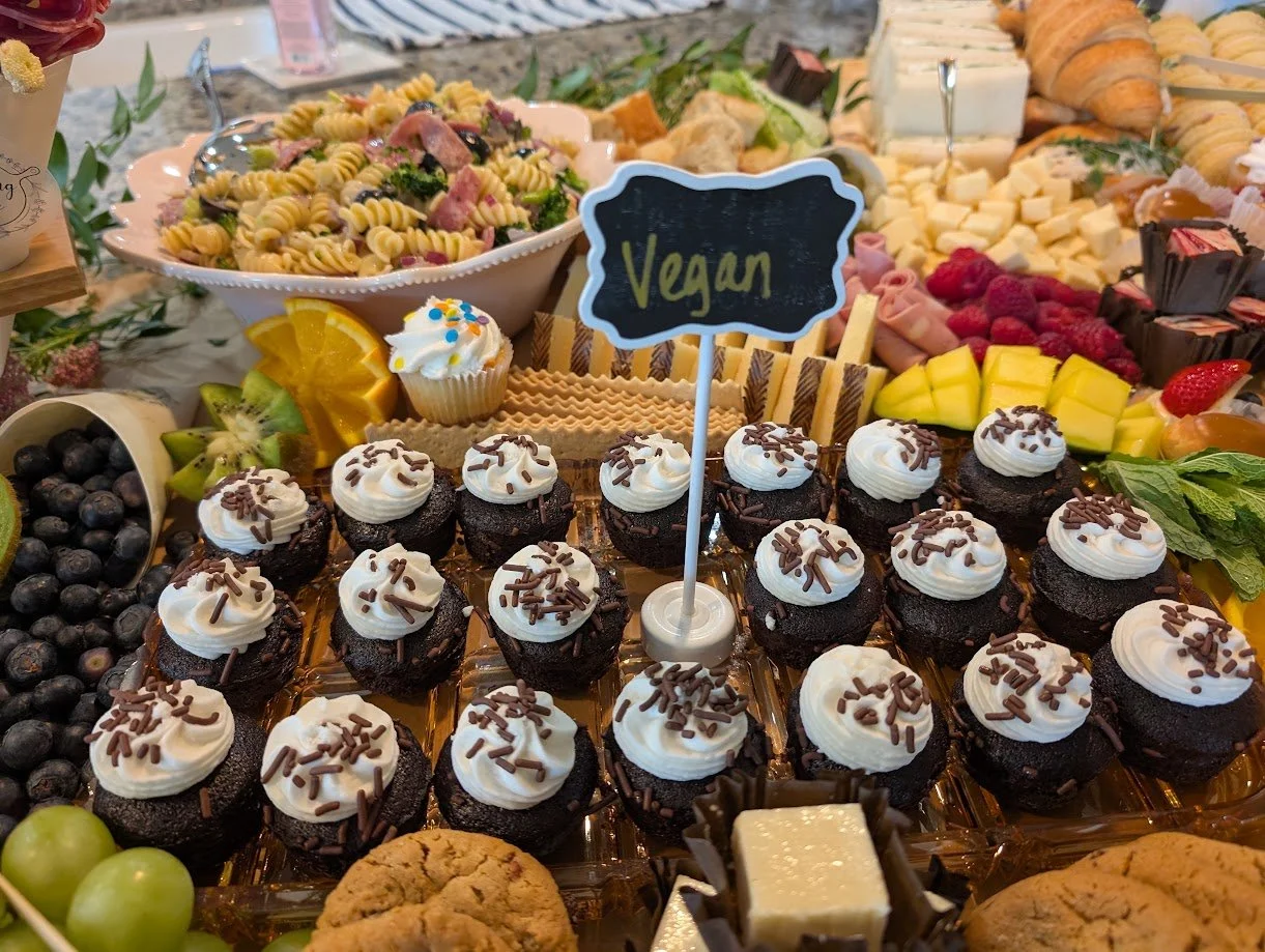 Vegan chocolate cupcakes with white frosting and sprinkles on a dessert table surrounded by various fruits, pasta salad, cheeses, and pastries with an upright sign reading 'Vegan'.