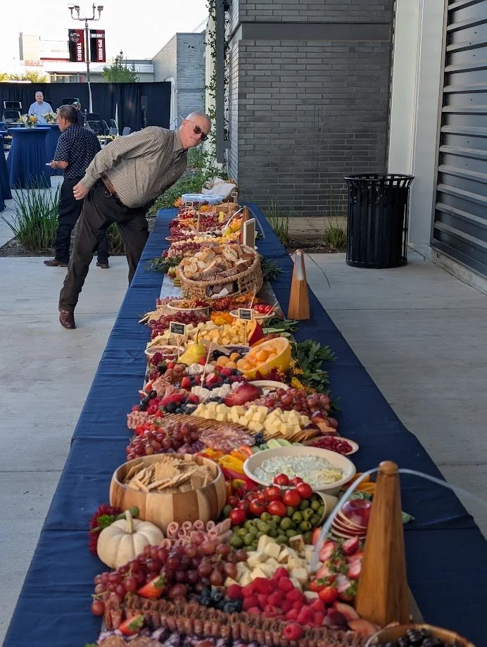 A long buffet table with various fruits, cheeses, and snacks, set up outdoors with people serving themselves.