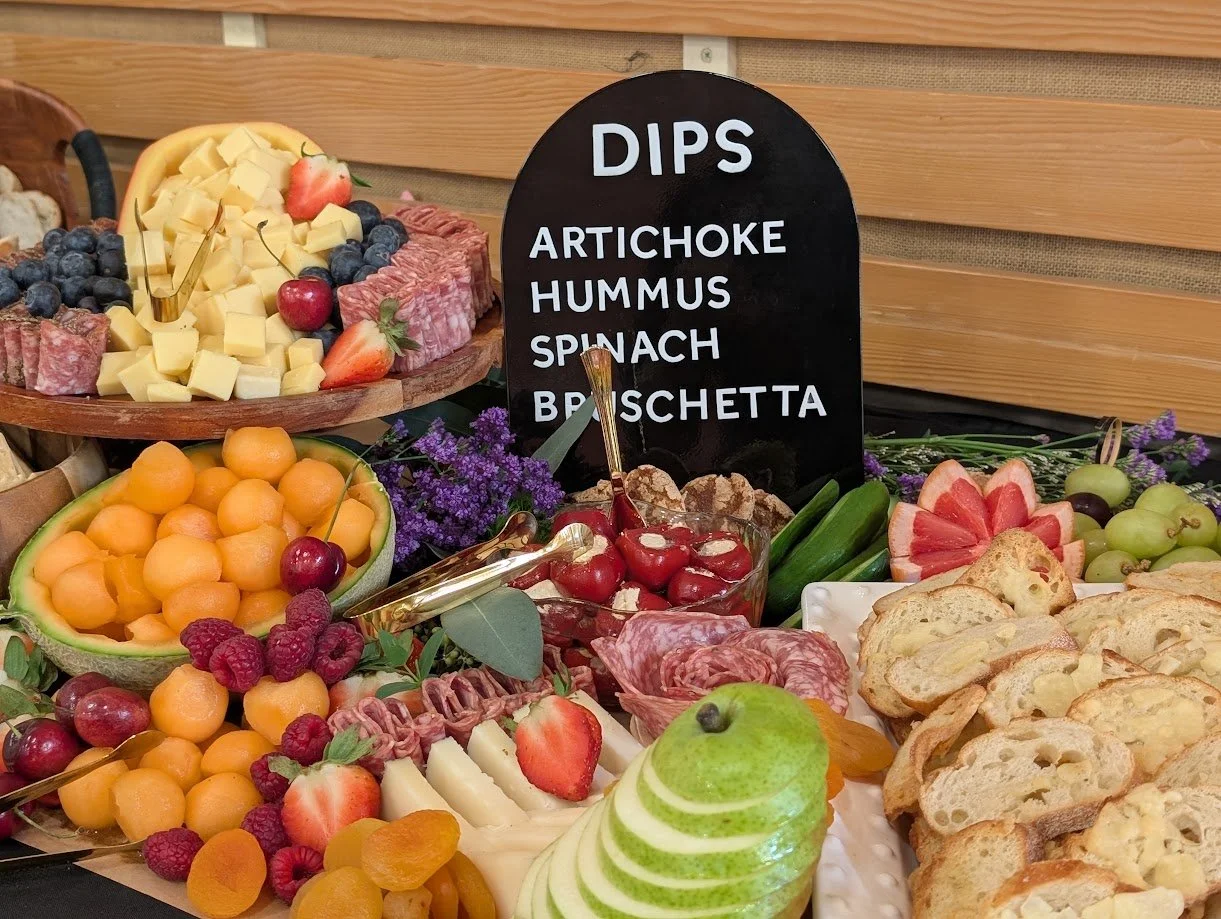 Assorted fruit, cheese, and charcuterie on a table with a blackboard sign listing dips: artichoke, hummus, spinach, bruschetta