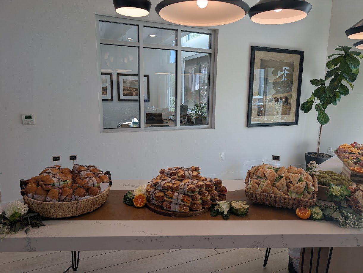 Table with sandwiches, burgers, and various snacks, decorated with flowers and fruits in a modern indoor setting.