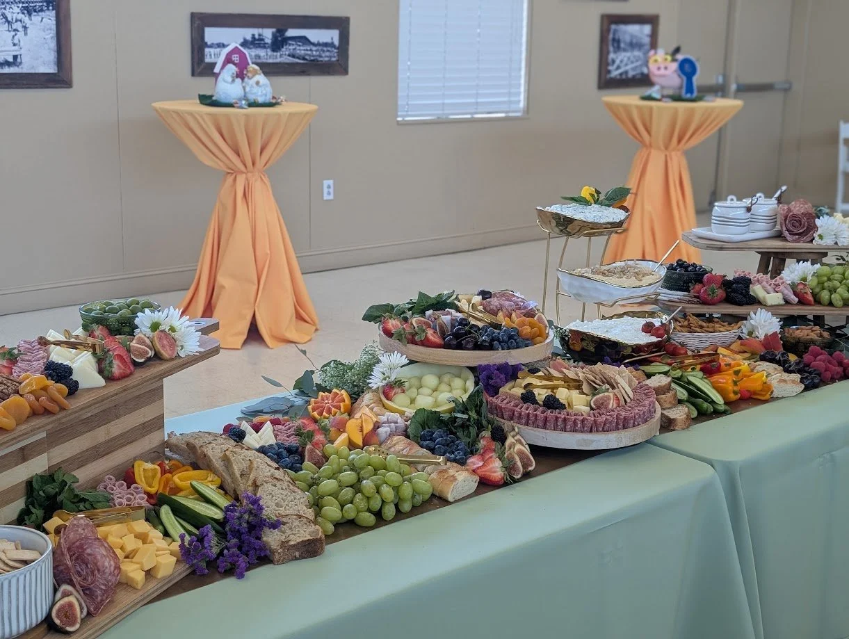 An elaborate buffet table with various cheeses, fresh fruits, vegetables, and appetizers, decorated with flowers, set for a gathering or celebration.