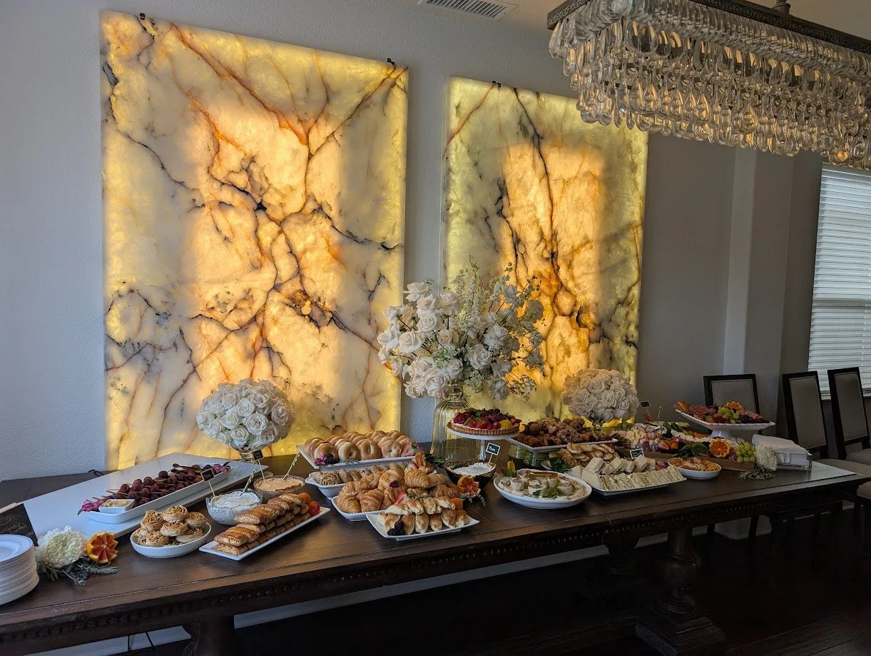 Buffet table with various pastries, fruits, and finger foods, decorated with white flowers, in front of backlit onyx wall panels and under a crystal chandelier.