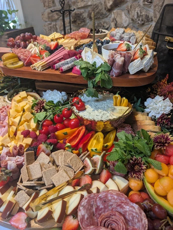 An elaborate charcuterie and cheese board with grapes, strawberries, cherry tomatoes, sliced meats, cheeses, crackers, colorful peppers, herbs, and various fruits and snacks arranged on wooden trays.