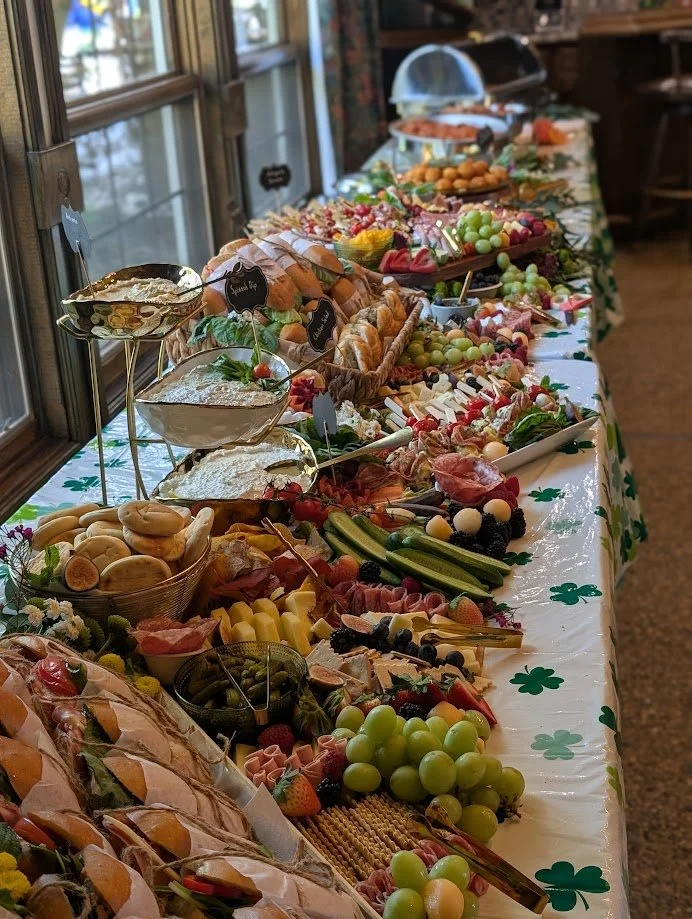 A long table covered with a green shamrock-themed tablecloth displays an assortment of finger foods, including sandwiches, fruits, cheeses, and desserts, set near a window with natural light.