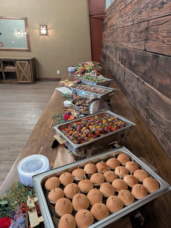 Buffet table with various food items including decorative buns, skewered vegetables, cooked meat, and desserts in a rustic setting.