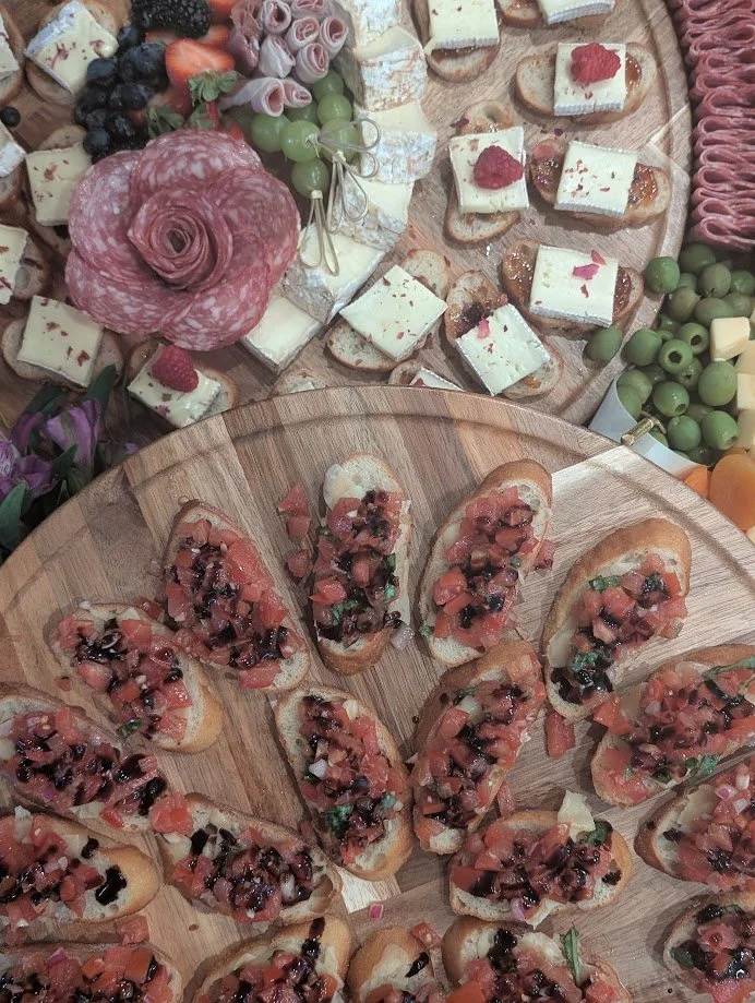 A wooden platter with sliced baguette topped with chopped tomatoes, herbs, and balsamic glaze, and another platter with assorted finger foods, cheese, fruit, and crackers.