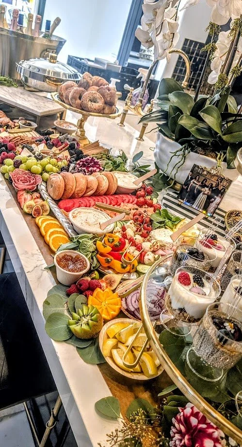 A lavish breakfast or brunch spread on a white marble table with a variety of fresh fruits, pastries, dips, and desserts, decorated with flowers and greenery.