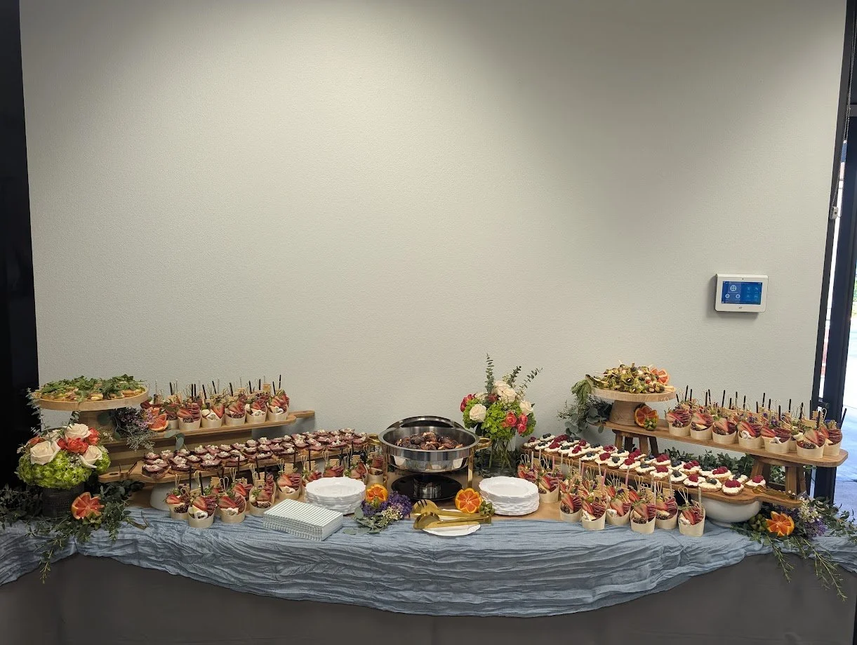 A buffet table with assorted appetizers and desserts, including small cups with skewers, cupcakes, and finger foods, decorated with flowers and greenery.