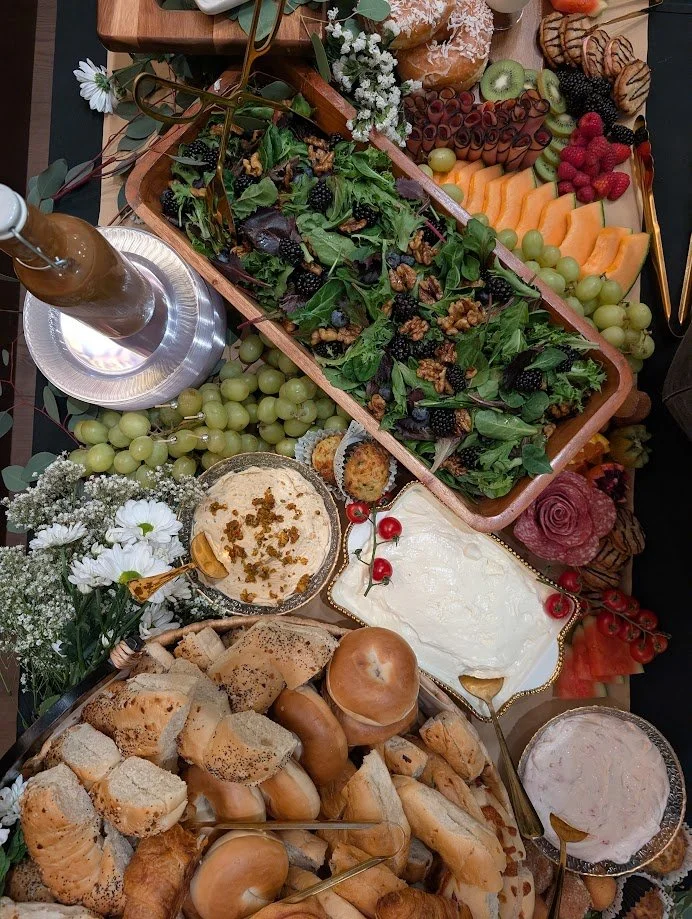 A table with a salad, assorted fruits including grapes, kiwi, strawberries, and melons, bread rolls, cheese, and desserts, decorated with flowers and greenery.