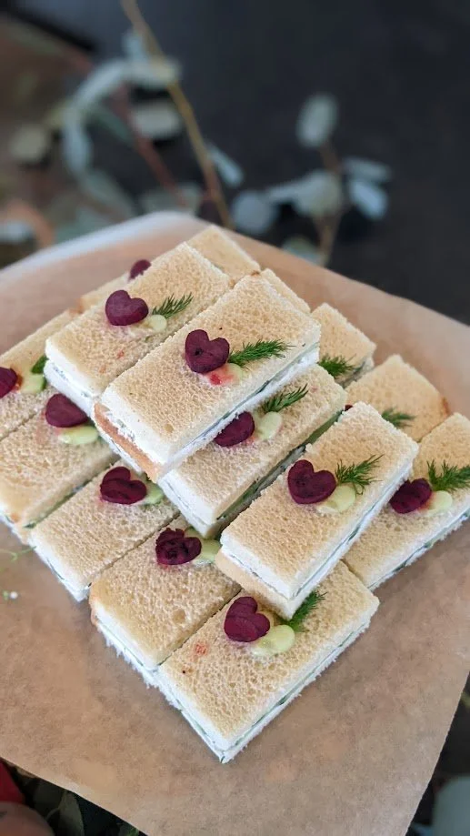 Assorted mini sandwiches with white bread, garnished with heart-shaped red toppings and small green herbs on a brown paper-lined tray.