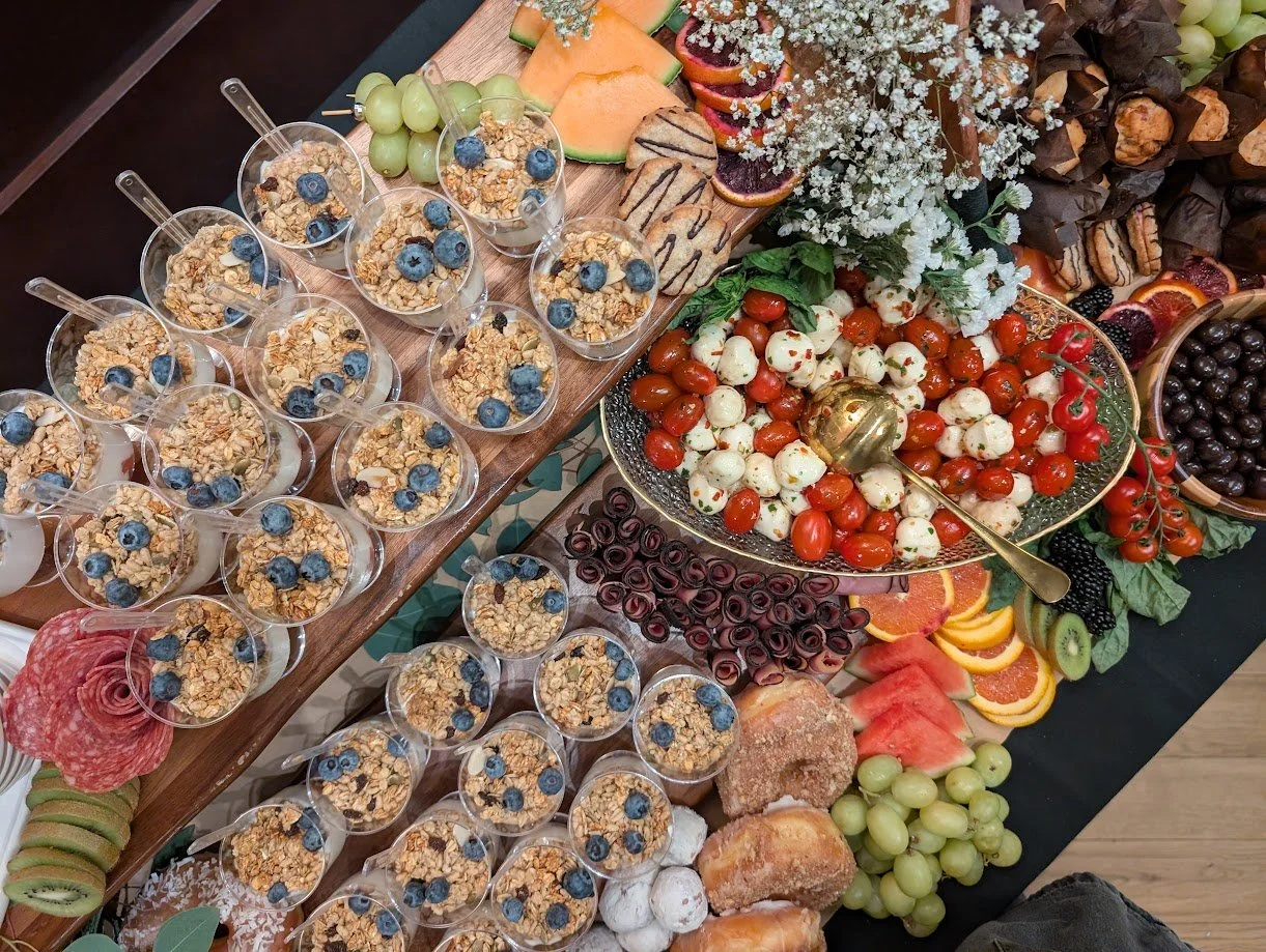 A large spread of assorted foods, including bowls of granola with blueberries, grape and melon slices, cherry and mozzarella skewers, assorted berries, watermelon, orange, kiwi, bread rolls, and appetizers, arranged on a table for a buffet or party.