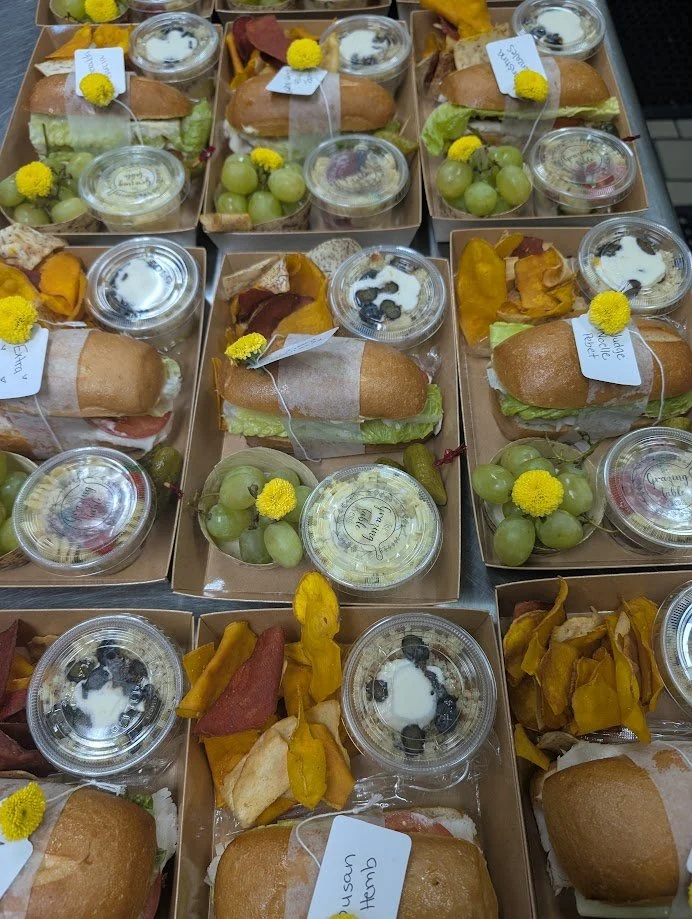 Multiple packed lunch boxes containing sandwiches, grapes, and small containers of yogurt or pudding with toppings, decorated with yellow flowers.