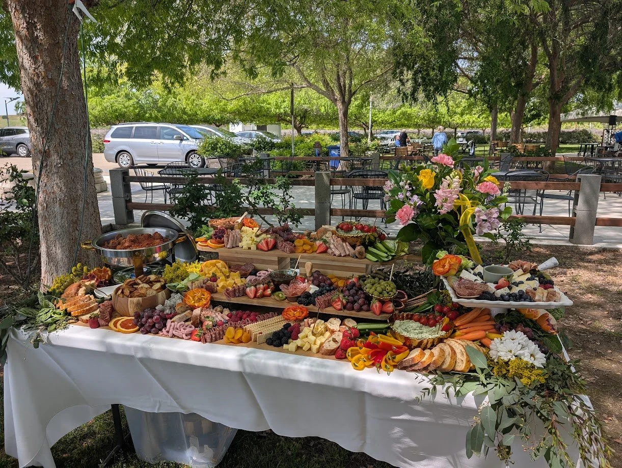 An outdoor buffet table with a variety of cheeses, fruits, vegetables, sliced bread, and a large flower arrangement, set under trees with a parking lot and people in the background.