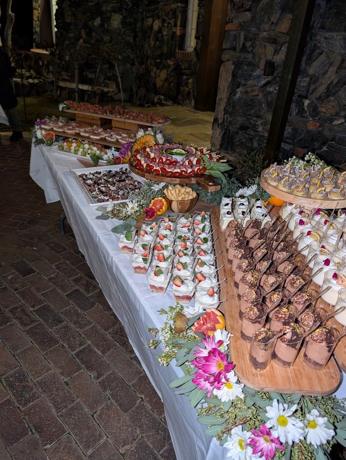 A dessert table at an event with various sweet treats, decorated with flowers and greenery. There are layered desserts with strawberries, chocolate mousse, and small pastry bites, arranged neatly on the table with decorative flowers along the edge.