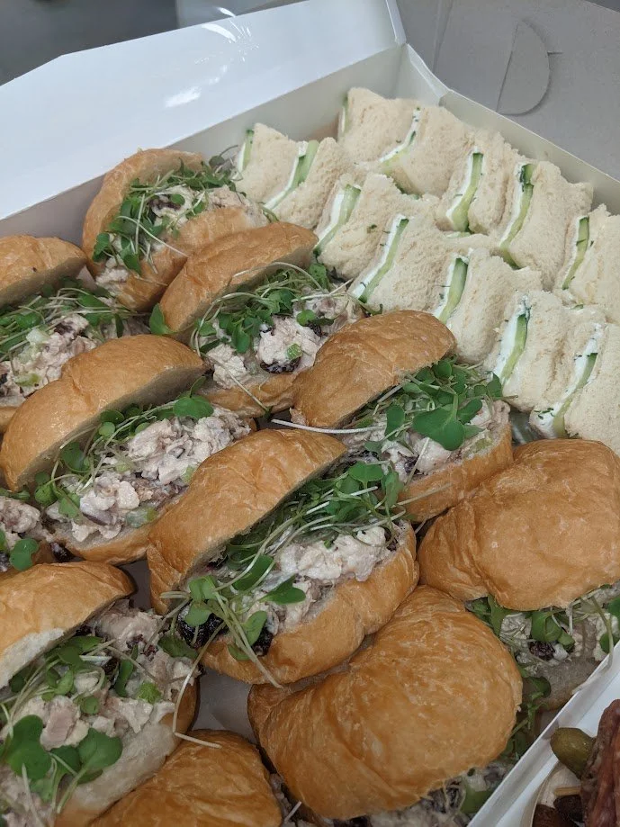 A box with sandwiches, including small sandwiches with cucumber slices and larger sandwiches filled with chicken salad topped with microgreens.